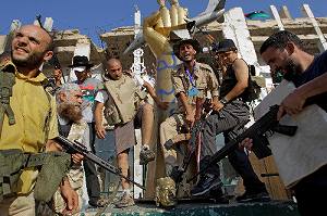 写真・図版 : トリポリのカダフィ政権軍の拠点、バーブ・アジジヤ地区に入り、カダフィ大佐の像の一部を踏みつけるしぐさをする反体制派の兵士たち＝８月23日、ＡＰ