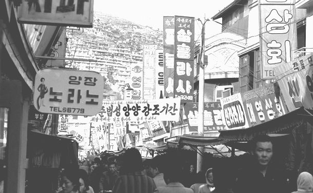 写真・図版 : 1965年当時の釜山国際市場。「商会」とか「洋装」のハングルが見えます。遠くの方に「遵法性昂揚強調期間」と横断幕があるのが分かりますか？