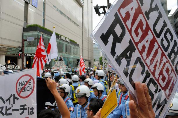 写真・図版 : 警察官に囲まれながら日の丸や旭日旗を手に歩くデモ隊（中央奥）に対し、ヘイトスピーチに反対するプラカードを掲げ、抗議の声を上げる人たち＝2016年6月、福岡市