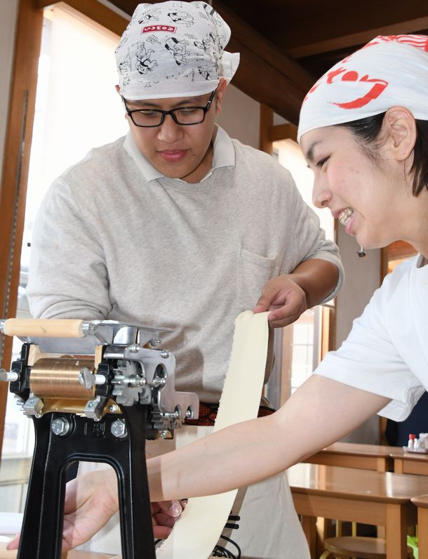 写真・図版 : ラーメンの生地を製麺機にかけるフィリピン人旅行者（左）と女性店員＝2018年6月14日、京都市東山区  
