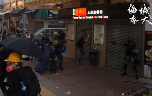 写真・図版 : 香港で1日、デモ隊に向けて銃を向けている警察官（右）に撃たれ、後ろに倒れようとする高校2年生の男子（中央、ピンク色のガスマスクをつけている人物）＝香港城市大学生会のフェイスブックの動画から 