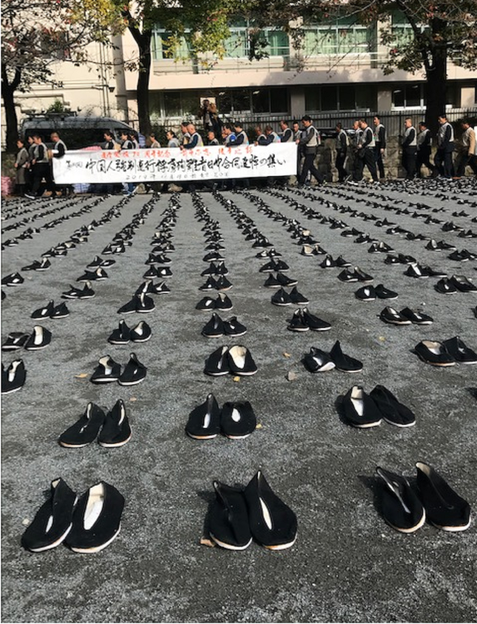 写真・図版 : 東京・芝公園に並べられた6830足の黒い革靴の後ろを通るデモの行列＝2019年11月19日、筆者撮影