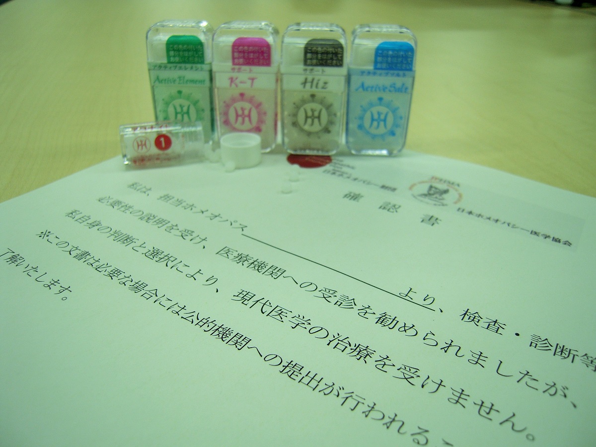 写真・図版 : 日本ホメオパシー医学協会が作成した利用者向けの確認書。「私自身の判断と選択により、現代医学の治療を受けません」と書かれ、利用者がサインする欄がある。奥は療法で用いられる砂糖玉