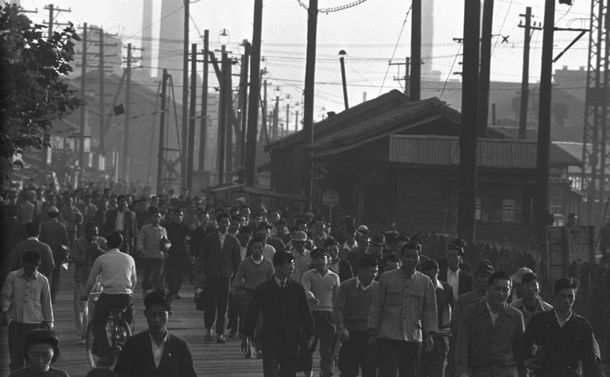 写真・図版 : 1950年6月25日に始まった朝鮮戦争を契機に日本の産業界は活況を呈した。戦争による「特需景気」の到来である。日本重工業地帯の一つ、京浜工業地帯の川崎・鶴見地区で大小の工場から黒煙が上がるなか、労働者たちが工場の門に向かう＝1950年10月、神奈川県川崎市 
