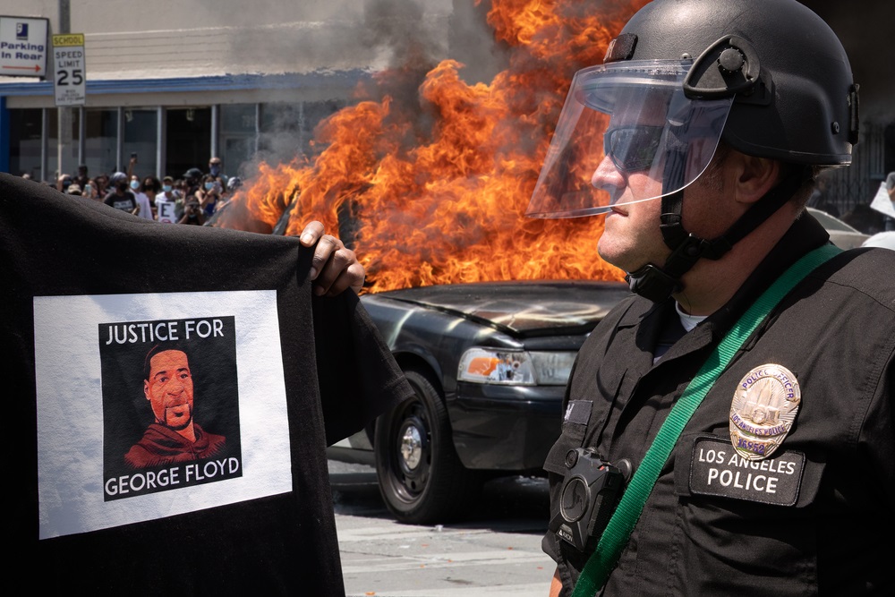 写真・図版 : 4人の警官に殺されたジョージ・フロイドさんの顔が描かれたTシャツを警官に向けて掲げ抗議する人＝2020年5月30日、米ロサンゼルスHayk_Shalunts / Shutterstock.com