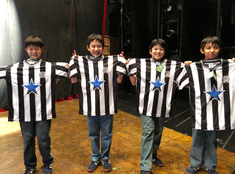 写真・図版 : 初⽇のお祝いに演出補のサイモンから主役の4⼈に、ビリーが好きなサッカーチームのユニホームが贈られた