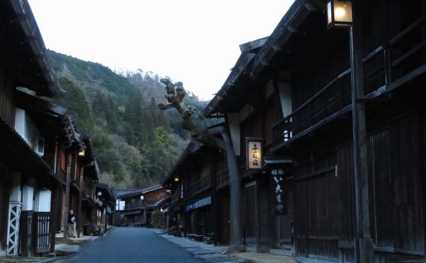 写真・図版 : 妻籠宿の寺下地区。アオギリの木とかつての宿屋などが軒を連ね、街道は正面の山々に吸い込まれるように延びる