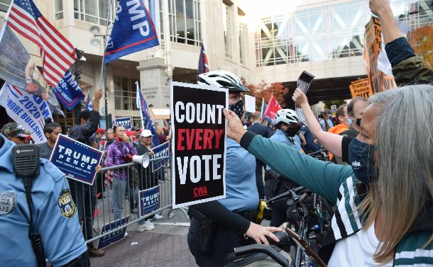 写真・図版 : 大統領選の開票作業が続く米ペンシルベニア州フィラデルフィアで、警官隊を挟んでトランプ大統領の支持者とにらみ合い、気勢を上げるバイデン氏の支持者ら（右）＝2020年11月6日、渡辺丘撮影