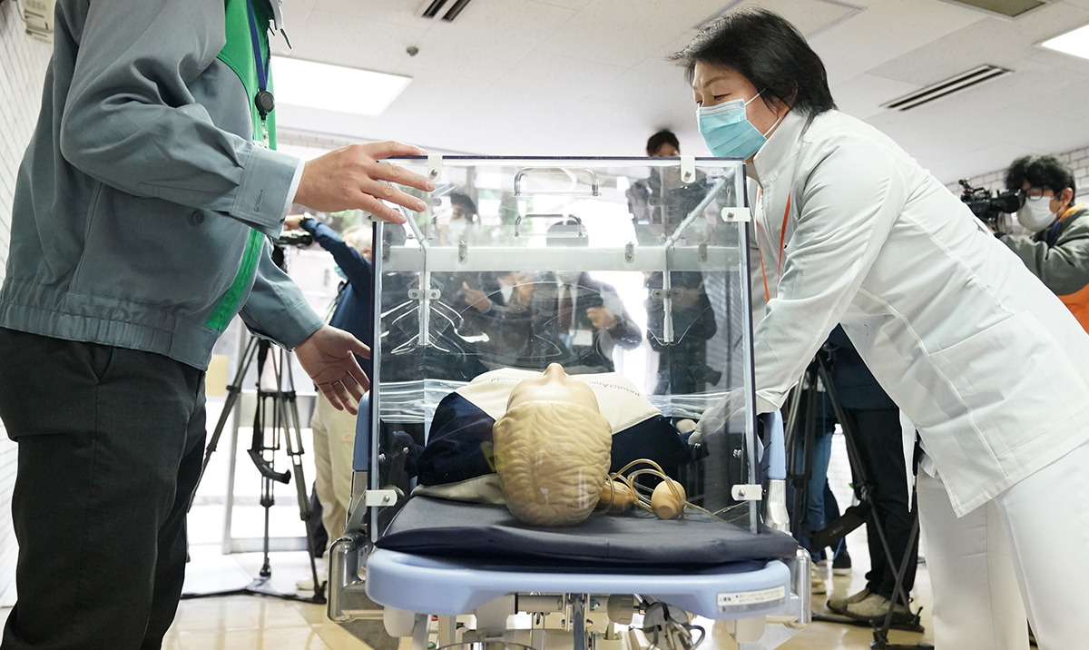 写真・図版 : 新型コロナ患者を想定し、アクリル製のカバーで覆ったストレッチャーを使って人形を運ぶ様子＝2020年12月2日、東京都府中市