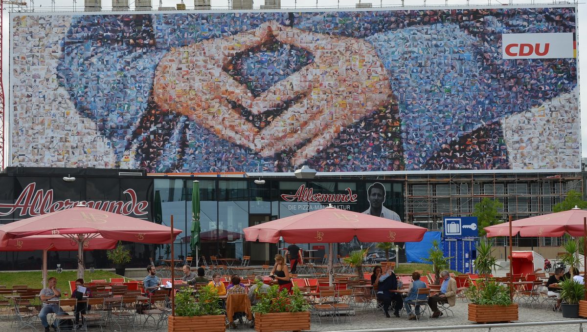 写真・図版 : トレードマークとなった「メルケルのひし形」。胸の下に指先が触れ合うように手を置く。キリスト教民主同盟はメルケル氏の強いリーダーシップの象徴としても利用。2013年の連邦選挙戦終盤にはベルリン中央駅前に巨大横断幕を設けた