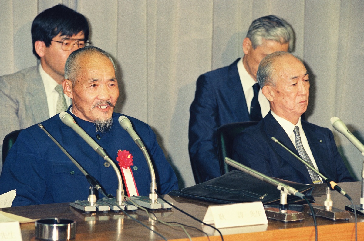 写真・図版 : 鹿島建設は1990年に花岡事件について企業の責任を認めて謝罪。同事件の生存者である耿諄さん（左）と、当時の村上光春副社長（右）がそろって会見した。耿諄さんの後ろが内田弁護士＝1990年7月5日、東京・鹿島建設本社
