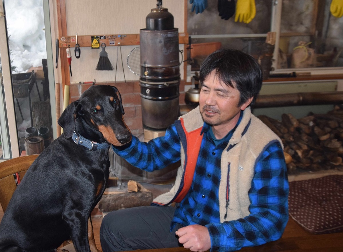 写真・図版 : 愛犬ベルをなでる木村紀夫さん。薪を燃やすロケットストーブで暖を取り、できるだけ電気は使わない=2015年3月、長野県白馬村北城