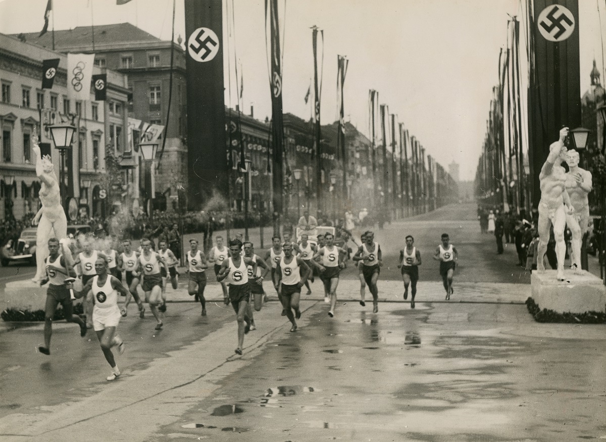 写真・図版 : オリンピック・スタジアムを目指してベルリンを走る聖火ランナー。五輪旗や立像、林立するナチス・ドイツのハーケンクロイツ旗で通りが飾られている＝1936年8月1日