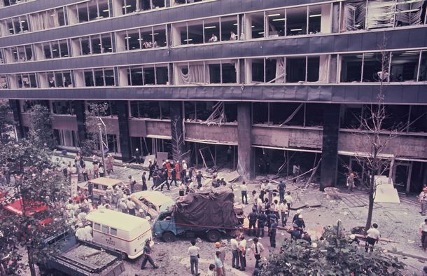 写真・図版 : ロビー1階に仕掛けられた爆弾が爆発し、建物9階まですべての窓ガラスが割れて地上に落下した三菱重工本社ビルの正面。割れた窓から社員たちが下をのぞきこみ、駆けつけた警察官らが上を見上げている。この爆発で同社の社員や通行人ら8人が死亡、380人が重軽傷を負った＝1974年8月30日、東京都千代田区丸の内2丁目