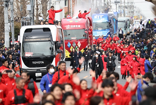 写真・図版 : 聖火リレーのリハーサルで、ランナー通過前に沿道の人たちを盛り上げるスポンサーの関係者と車両＝2020年2月15日、東京都羽村市