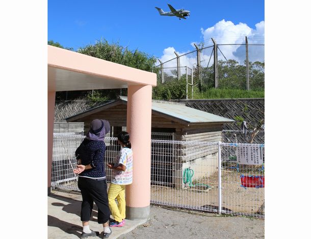 写真・図版 : 普天間飛行場に隣接する普天間第二小学校には2017年12月、米軍ヘリの窓が体育の授業が行われていた校庭に落下した。翌年に沖縄防衛局が建てた避難施設（左）は児童約100人が逃げ込める。その近くを普天間飛行場に着陸する米軍機が飛ぶ＝2018年9月、沖縄県宜野湾市