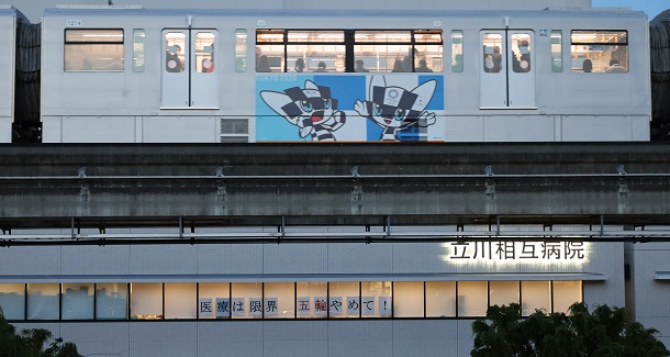 「医療は限界　五輪やめて！」とメッセージが貼り出された病院の横を、東京五輪のマスコットが描かれたモノレールが通過していった＝２０２１年５月６日午後６時４３分、東京都立川市、嶋田達也撮影 20210506