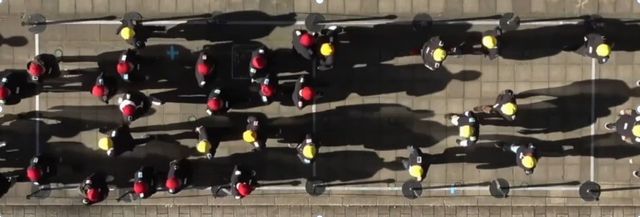 写真・図版 : 図1：実験を上から見た様子。赤の帽子の27人は右へ、黄色の帽子の27人は左に歩く（通路幅は3メートル）。この実験では、真ん中付近で、歩きスマホをしている赤い帽子の人と、黄色い帽子の対向者がぶつかっているのが分かる。