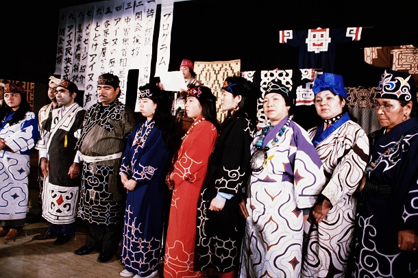写真・図版 : 中曽根康弘首相の単一民族国家発言に対して、「日本は単一民族の国ではありません」のスローガンを掲げた「アイヌ民族が存在することを、アイヌ自らがアピールする東京集会」が1986年11月30日、東京都品川区で開かれた。アイヌ民族の参加者のほとんどは民族衣装の正装。整列して集会声明を採択した 
