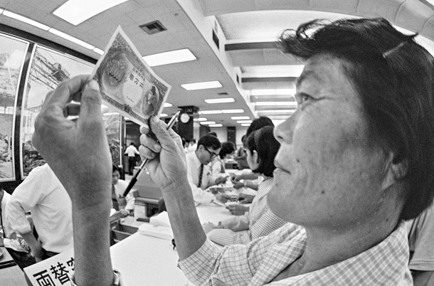 写真・図版 : 那覇市の琉球銀行で使い慣れた米ドルを日本円に交換し、見慣れない1万円札をじっと見る女性=1972年5月15日