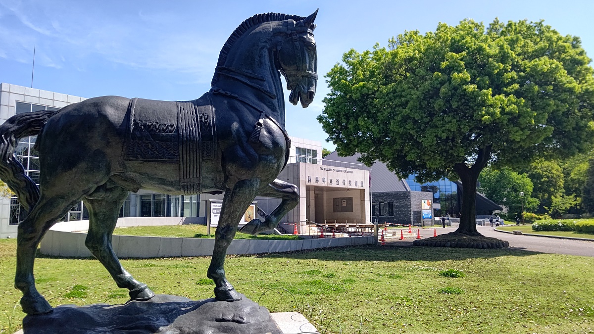 写真・図版 : 高さ4.5メートルのブロンズ像「巨きな馬」。奥に群馬県立の近代美術館と歴史博物館が並ぶ＝群馬県高崎市、筆者撮影