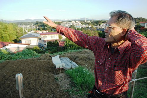 写真・図版 : 自家菜園の前に立つ荒尾さん。奥に見える阿武隈山系の向こうに福島第一原発がある
